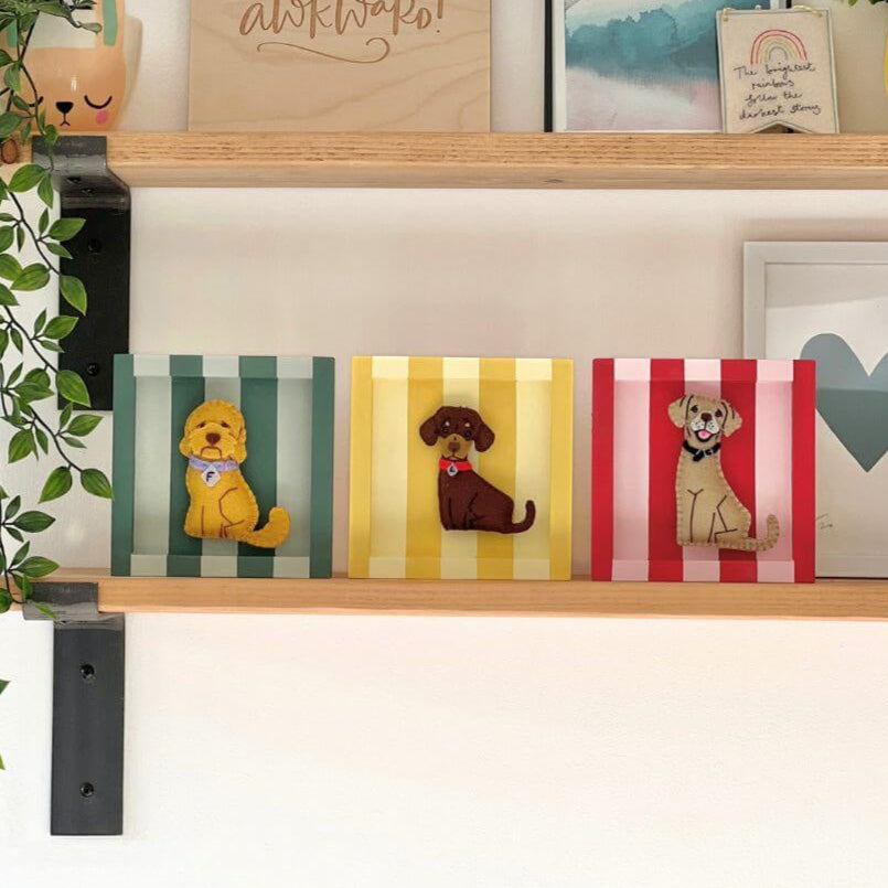 Three striped frames, one green, one yellow and the last one red and pink. Each had a felt dog ornament displayed inside. One is an Australian labradoodle, a sausage dog and a golden labrador. The sit on a shelf.