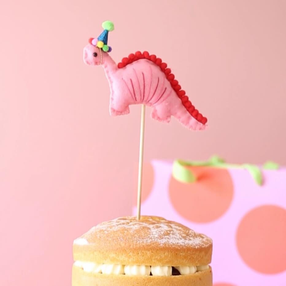 Pink dinosaur cake topper. The dinosaur wears a blue party hat with a green pom pom