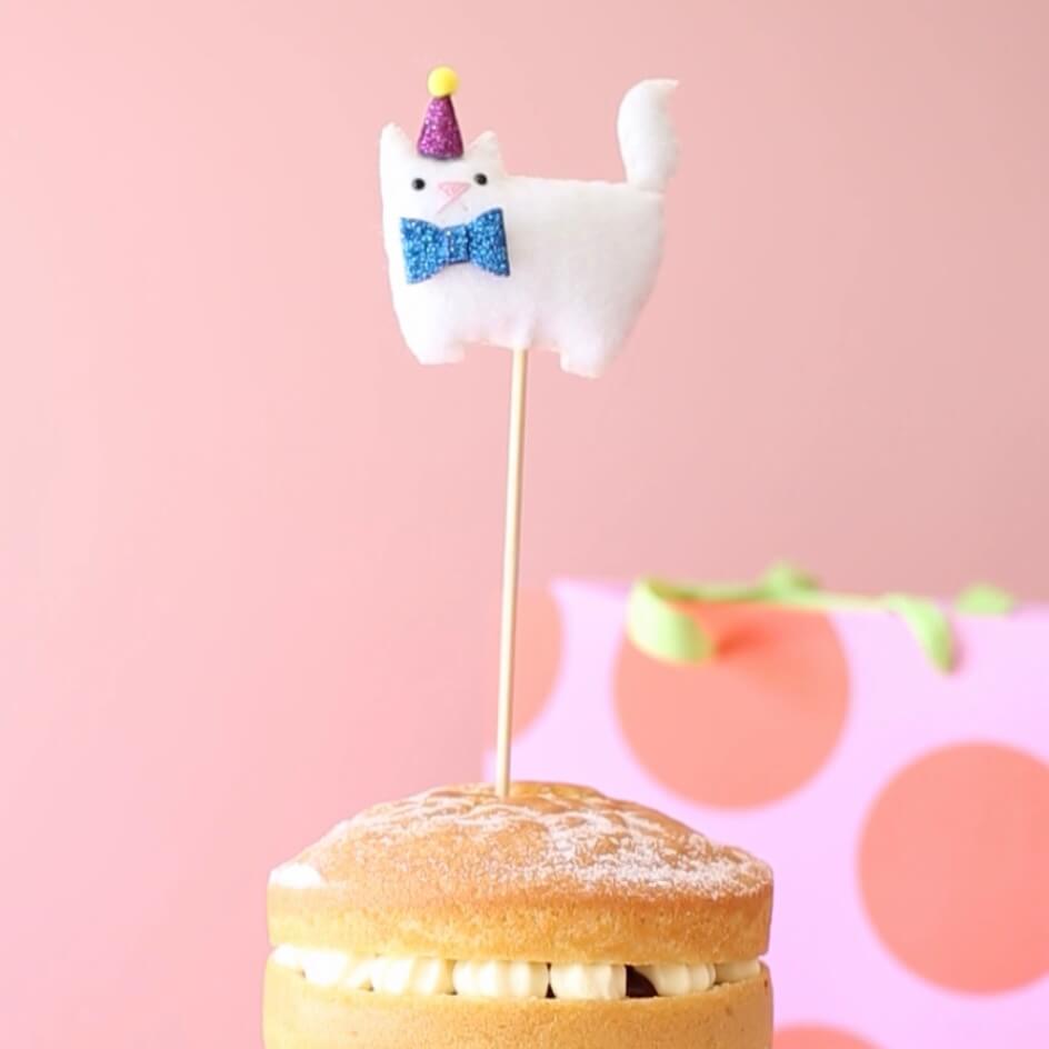 Cat birthday cake topper. A white cat with a purple party hat and a blue bow tie.