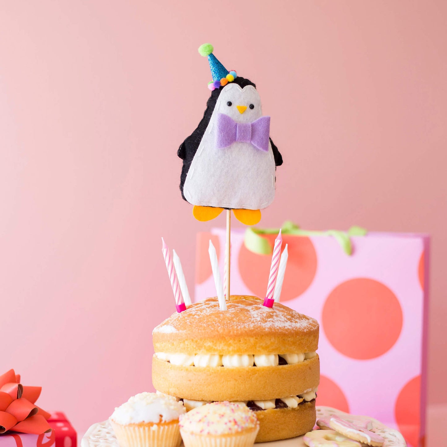 Felt penguin cake topper wearing blue pom pom party hat. He has a purple bowtie