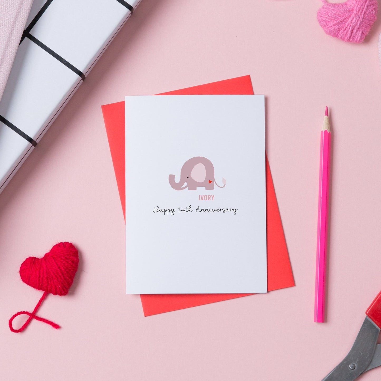 14th wedding anniversary card with a picture of an elephant on the front and text saying Happy 14th Anniversary. The card is on a pink background on top of a red envelope.