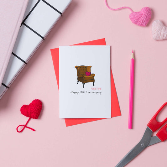 17th anniversary card with a picture of a leather armchair on the front. The card is placed on a red envelope and has some hearts, scissors and a pencil next to it.