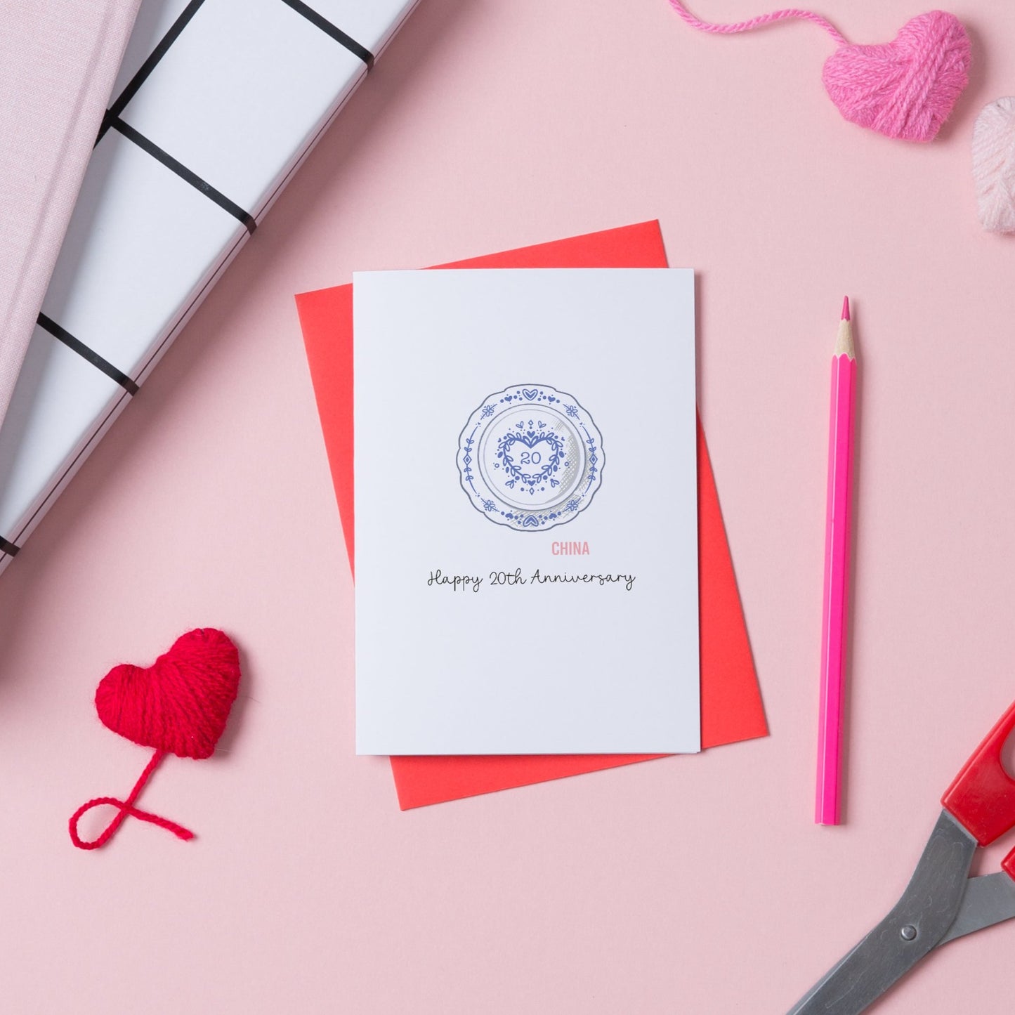 A white card with an illustration of a china plate with the number 20 in the middle. Underneath are the words Happy 20th Anniversary. The card is on top of a red envelope.