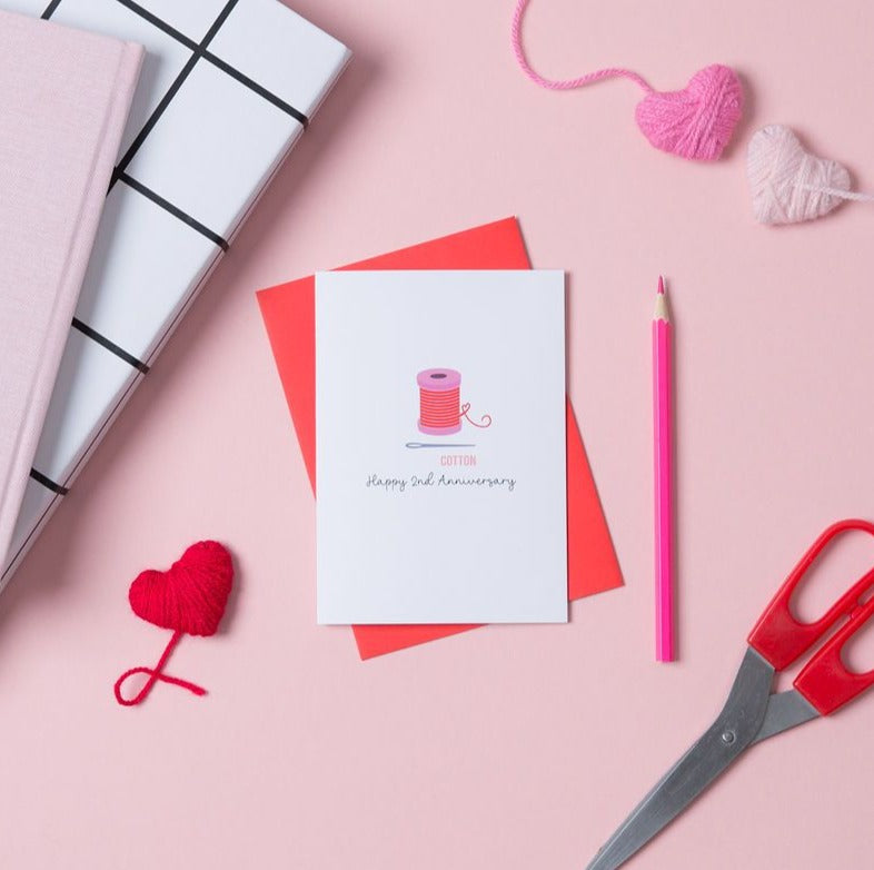 a card with a drawing of a cotton reel on the front. It says happy 2nd anniversary underneath. The card is on top of a red envelope.