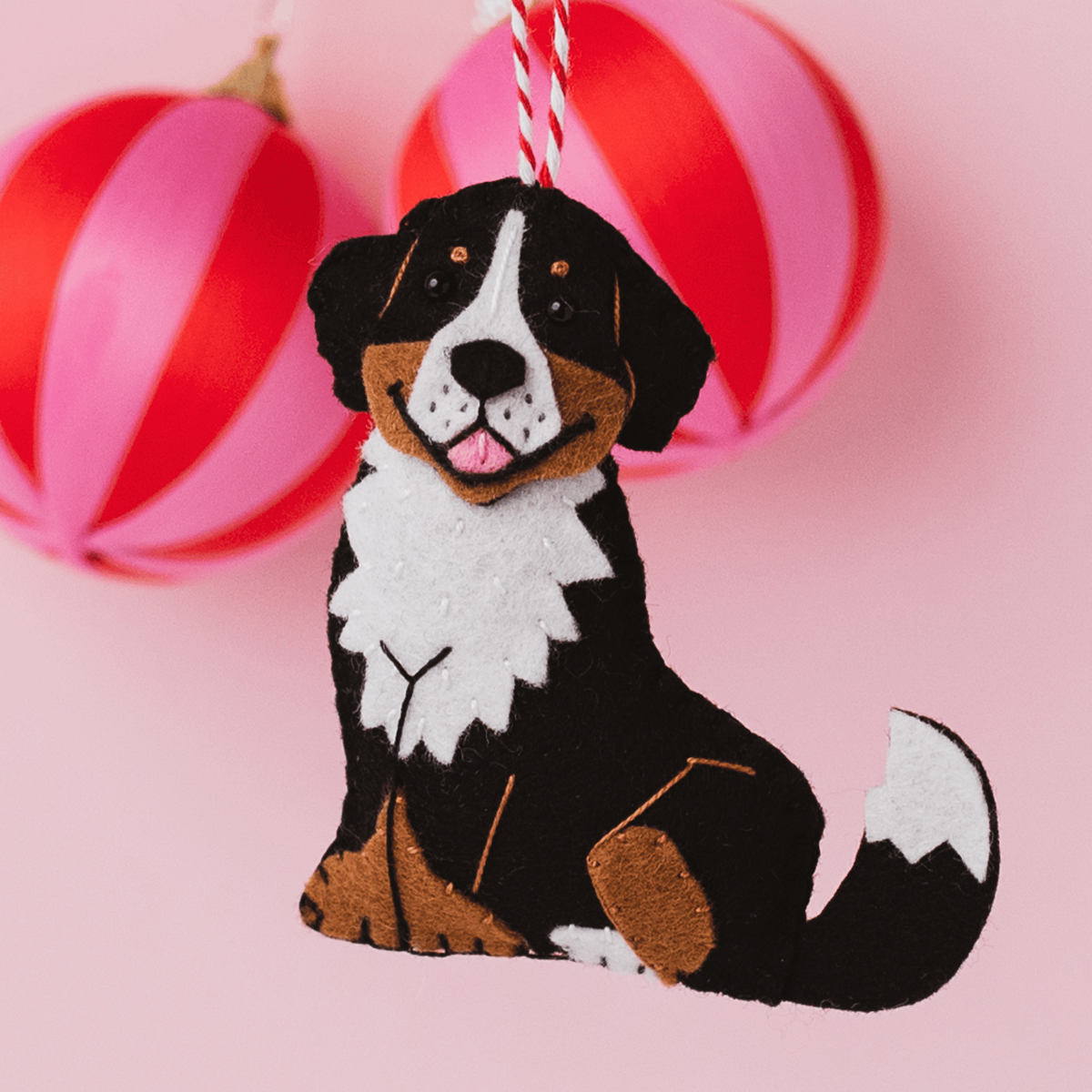 A felt dog ornament shaped like a Bernese mountain dog, hanging against a pink background with red-and-pink striped ornaments blurred behind it.