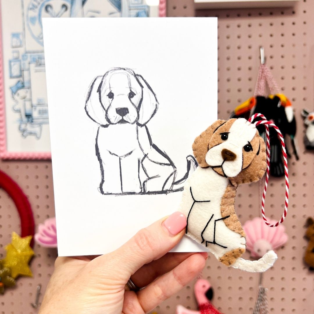 A hand holds a white card with a simple black line drawing of a dog next to a small handmade felt puppy ornament. 