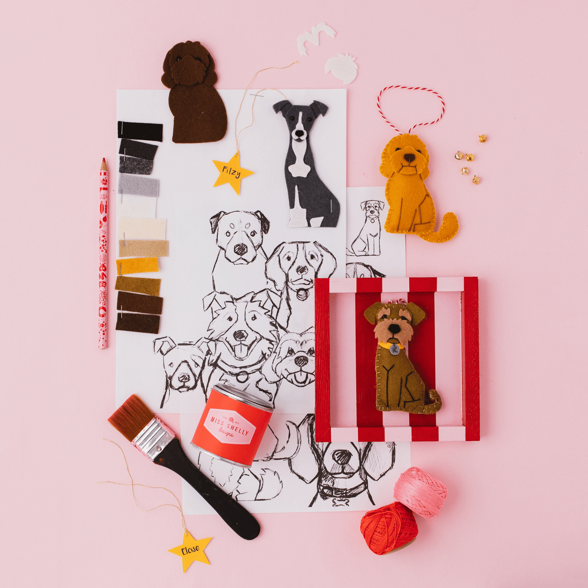A styled flat-lay of the design process of creating a bespoke pet doecoration, including felt dog cutouts in different breeds, a striped frame, threads, a paintbrush, colour swatches, and a sketch sheet with dog illustrations.
