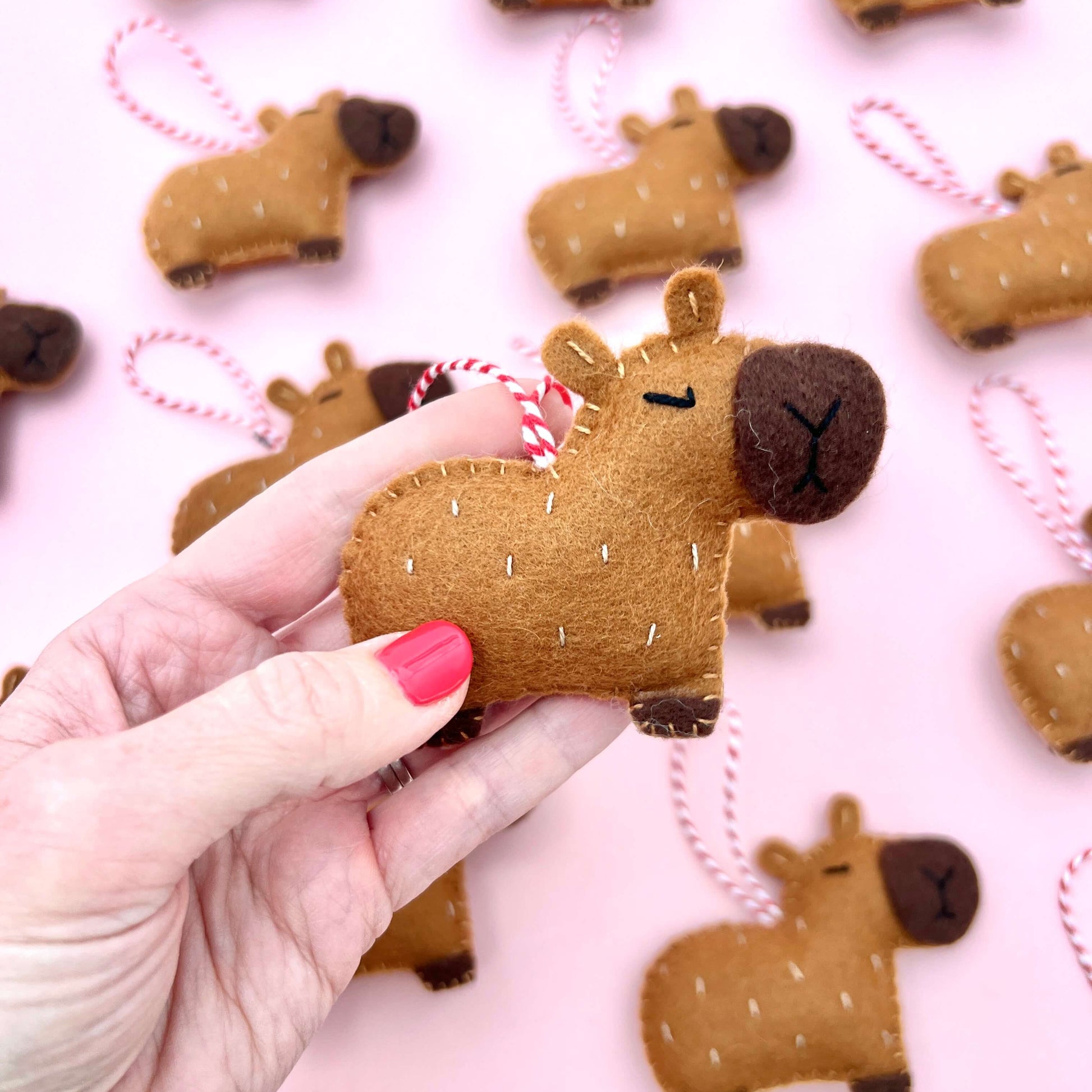 Hand holding a felt capybara ornament with more ornaments on a pink background