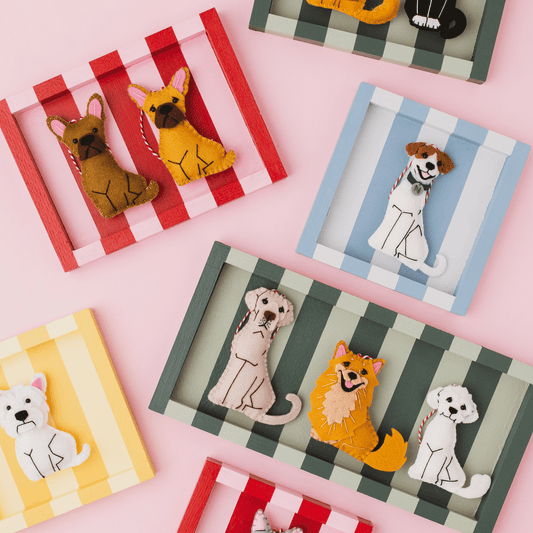 A styled flat-lay of multiple felt dog and cat fornaments displayed in striped wooden frames on a pink background.