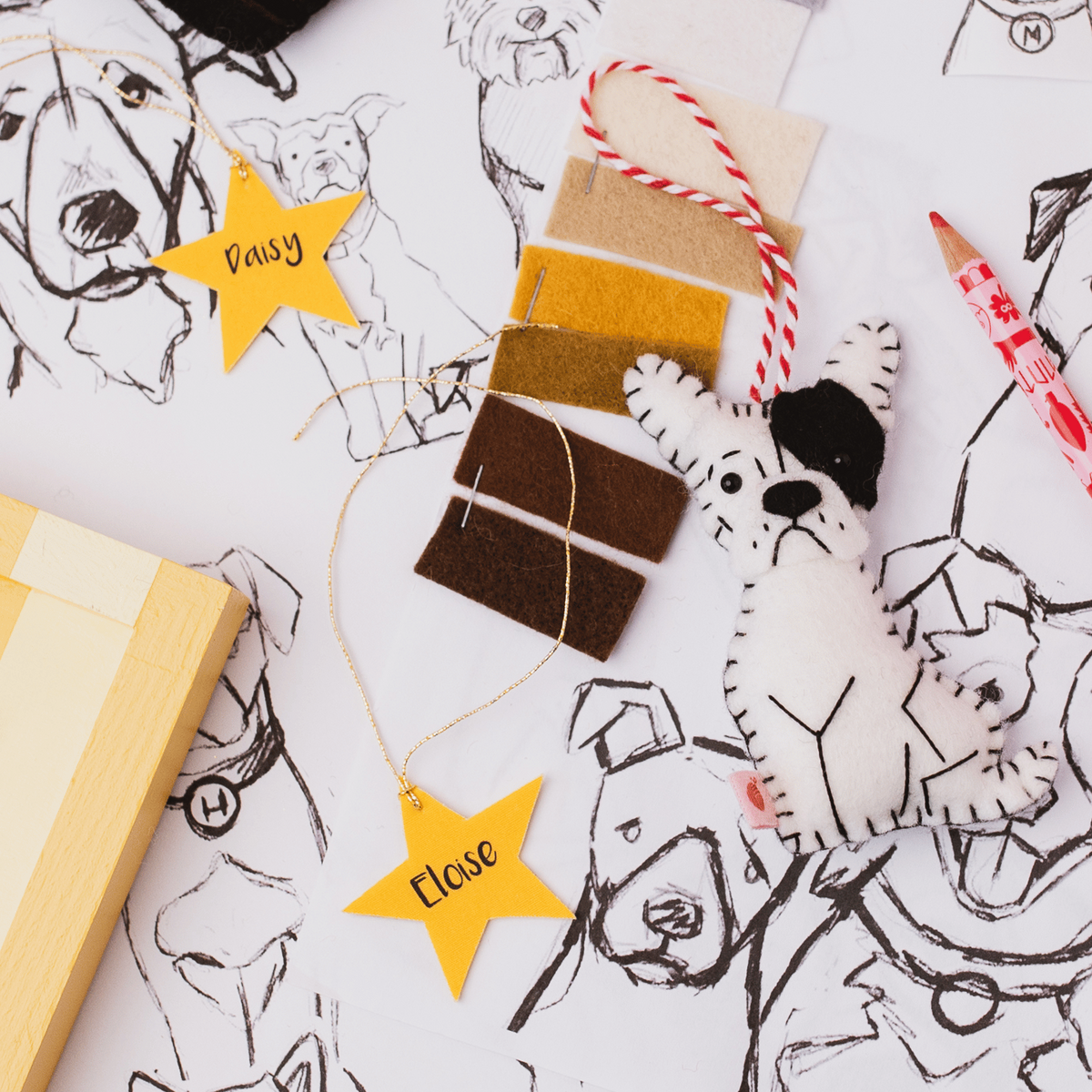 A styled flat-lay of dog-themed felt craft supplies on a white sketch pad. The set includes a white and black felt dog ornament, yellow felt star tags reading pet's names, brown and mustard felt swatches, a red-and-white striped hanging loop, and a wooden pencil.