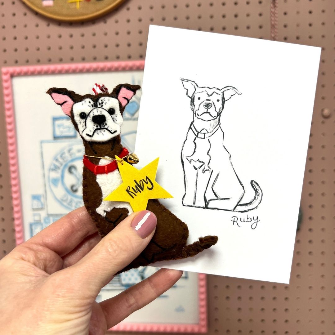 A hand holds a custom felt pet portrait of a brown and white dog with a red collar and a yellow star tag with the pet's name. Next to it is a matching black ink sketch on a white card.