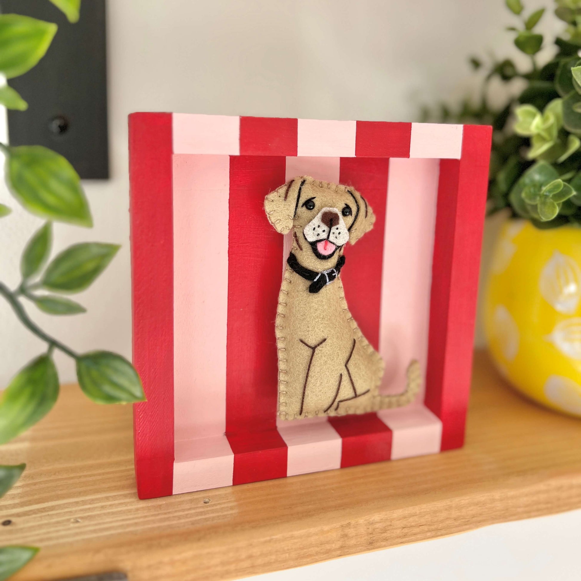 A golden labrador ornament in a red and pink striped frame. the frame is sat on a shelf next to plants.
