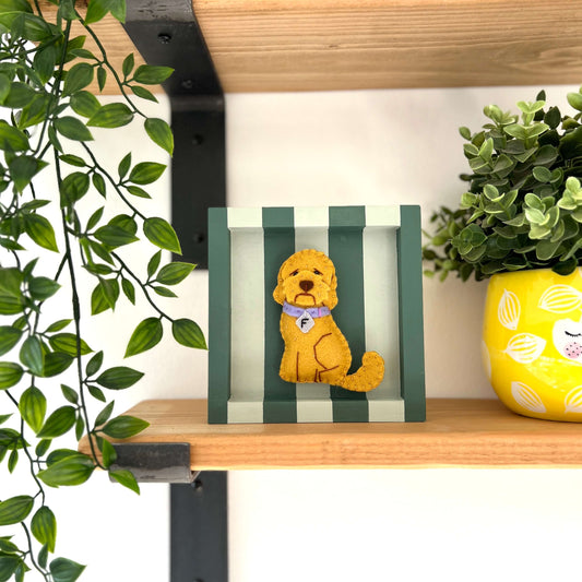 Labradoodle dog ornament in a golden colour felt. Placed in the center of a green striped box frame. Sitting on a shelf surrounded by green plants.