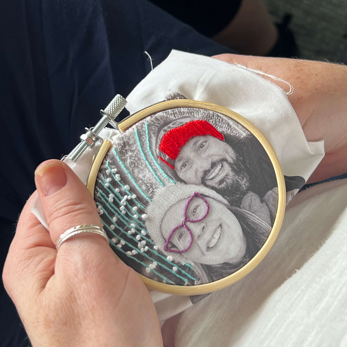 A close-up of a person holding a circular embroidery hoop with a custom portrait being stitched. The embroidery features a black and white photo of a couple with a bright red hat and purple glasses, set on white fabric.