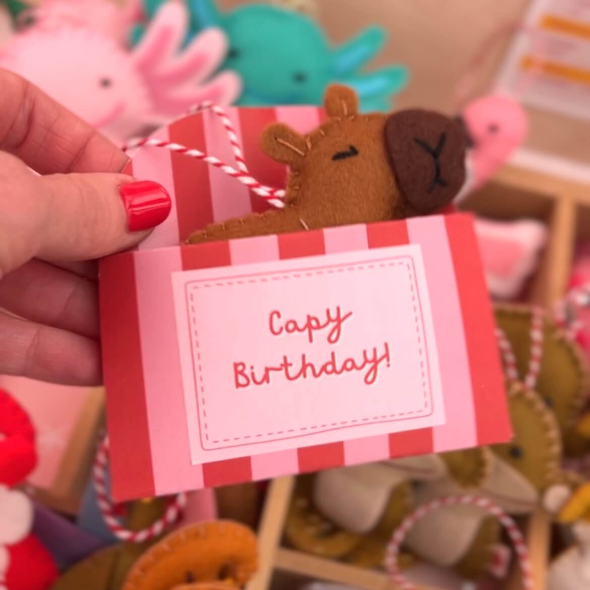 Small capybara ornament in a red and pink striped box with a 'Capy Birthday!' sticker, held by a hand.