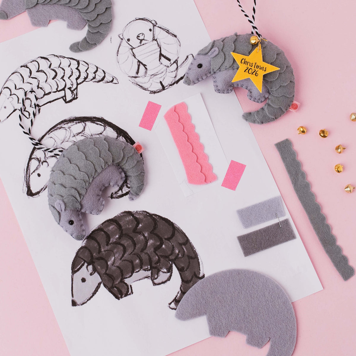 A flat-lay of a felt pangolin ornament on a pink background, showing grey felt pangolins in different sizes, black ink sketches, pink and gray felt strips, a yellow star tag reading 'Christmas 2026'.