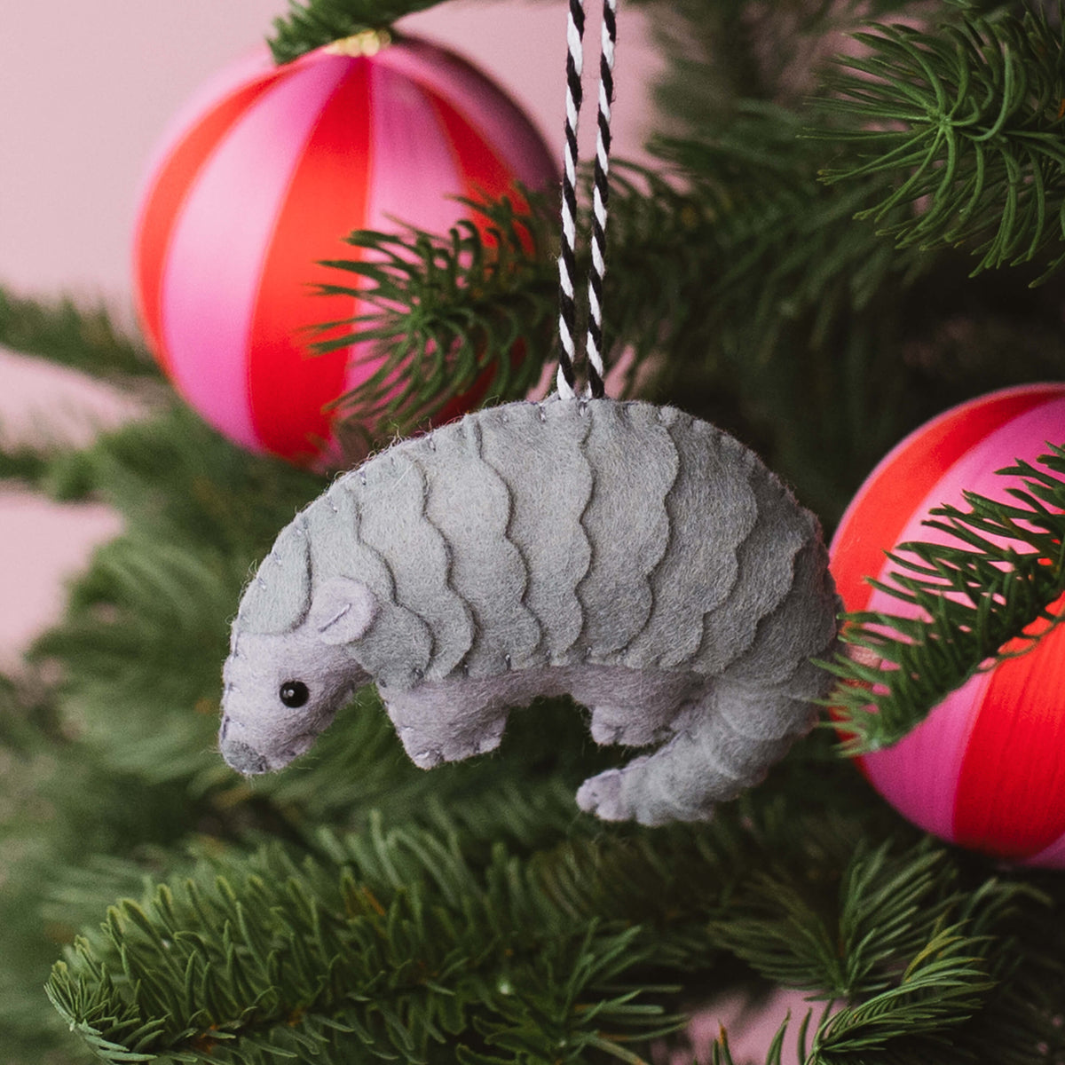 A gray felt pangolin ornament hanging from a black-and-white striped string among green pine branches, with pink and red striped Christmas ornaments in the background.