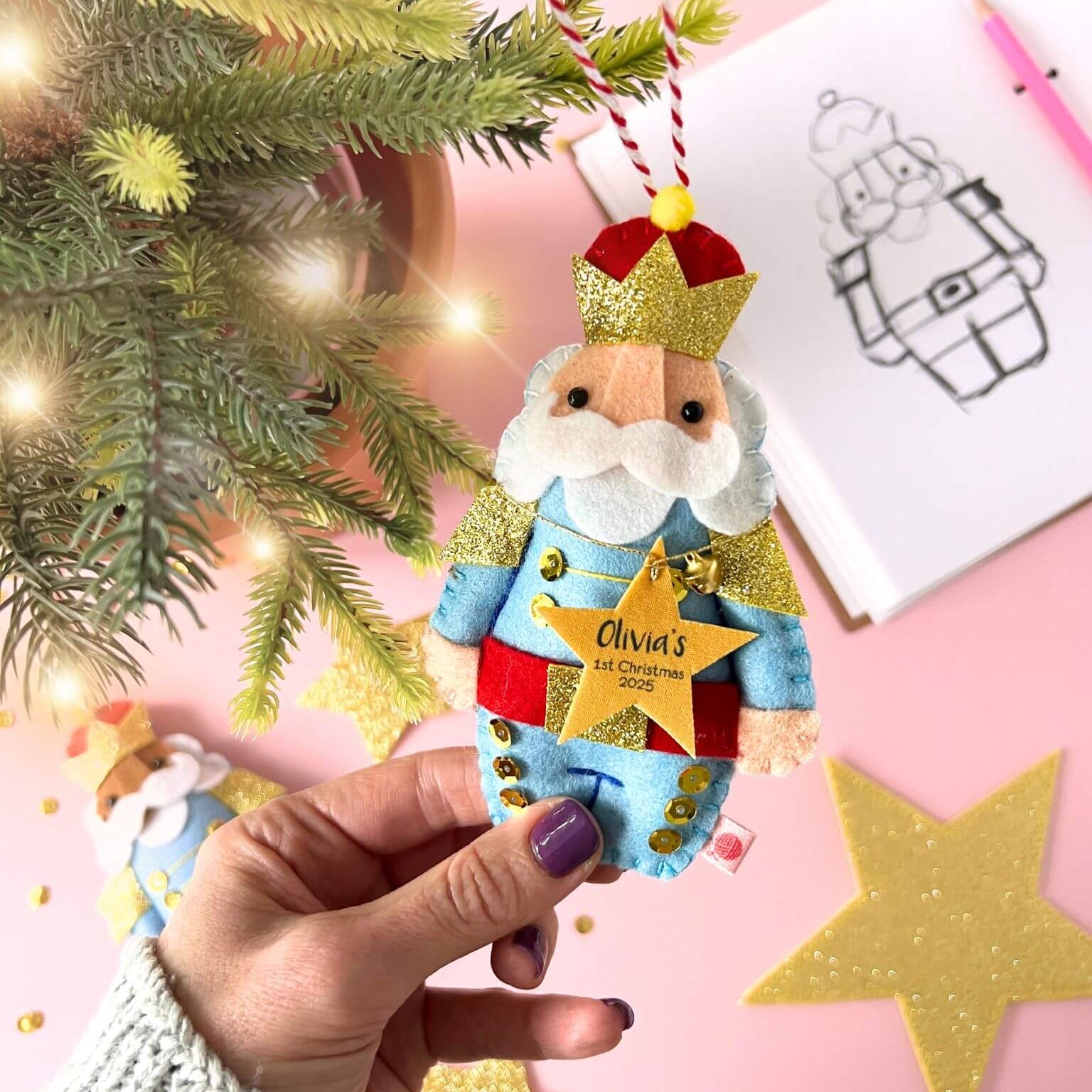 Hand holding a nutcracker Christmas ornament with a star saying 'Olivia's 1st Christmas 2025', against a pink background with a Christmas tree.