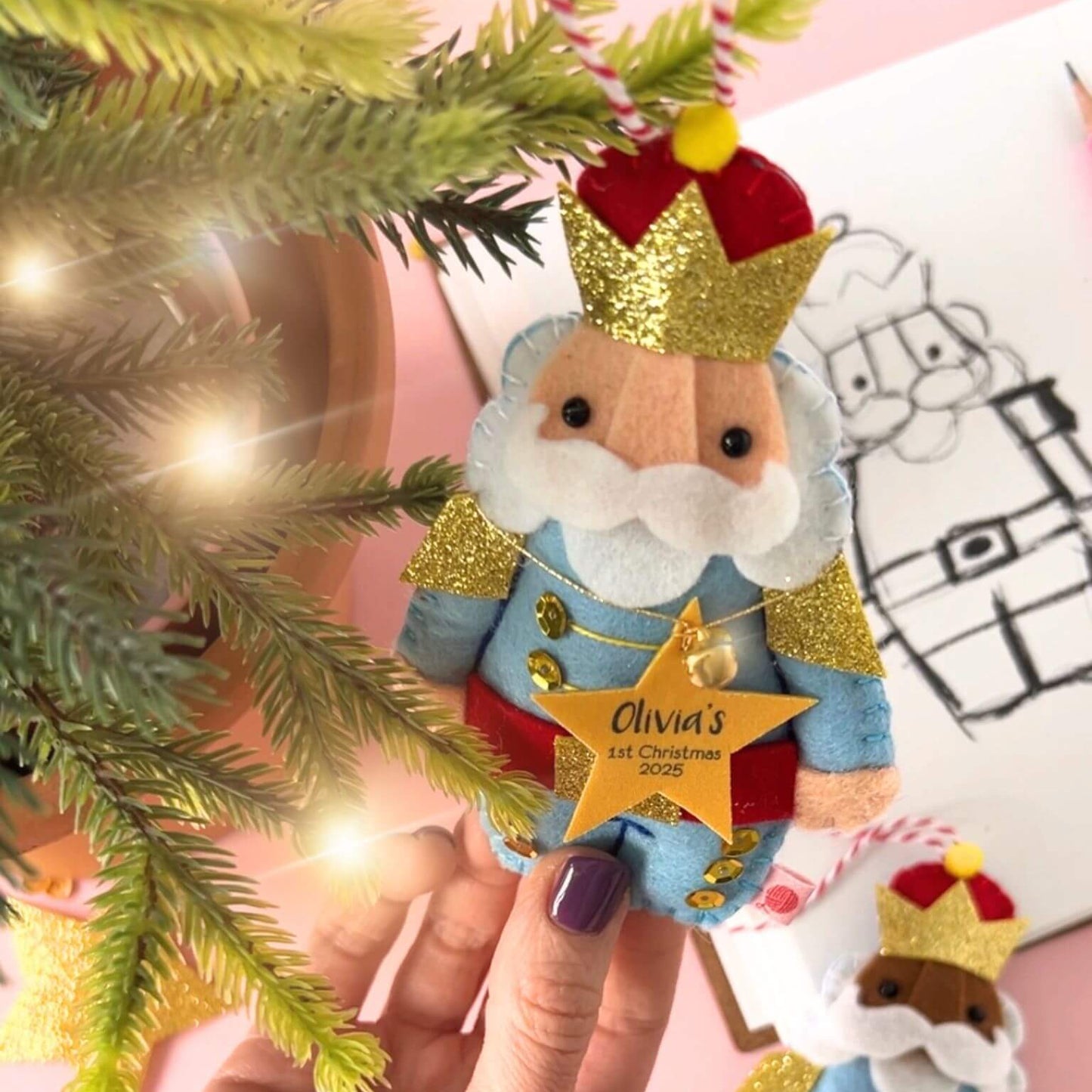 Felt Nutcracker Christmas Ornament being hung on a Christmas tree. There is a sketch of the Nutcracker in the background.