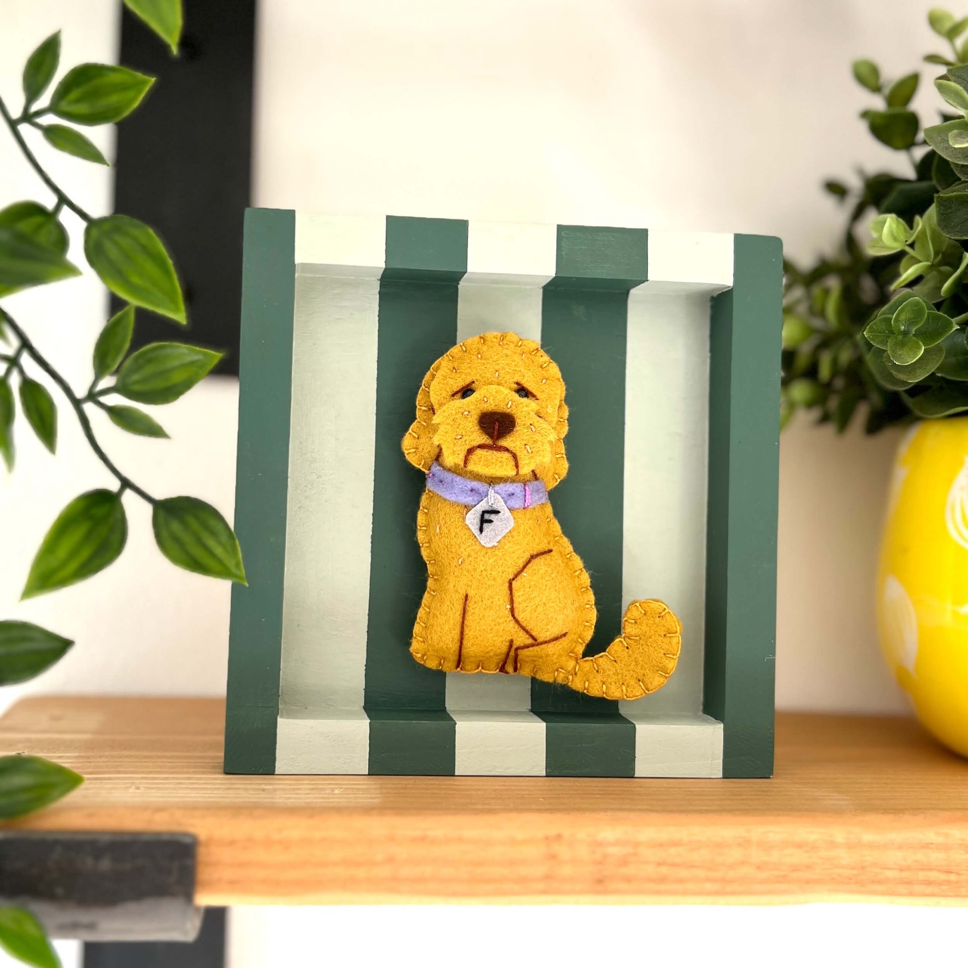 Labradoodle dog ornament in a golden colour felt. Placed in the center of a green striped box frame. Sitting on a shelf surrounded by green plants.