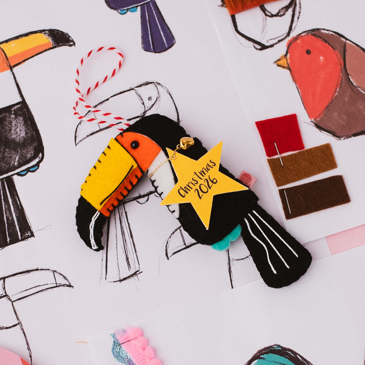 A handmade Christmas ornament shaped like a toucan, featuring black felt body, yellow and orange beak, red-and-white striped hanging string, and a yellow star tag that reads 'Christmas 2026'. The ornament is photographed from above on a white surface surrounded by bird illustrations, paintbrushes, and fabric swatches.