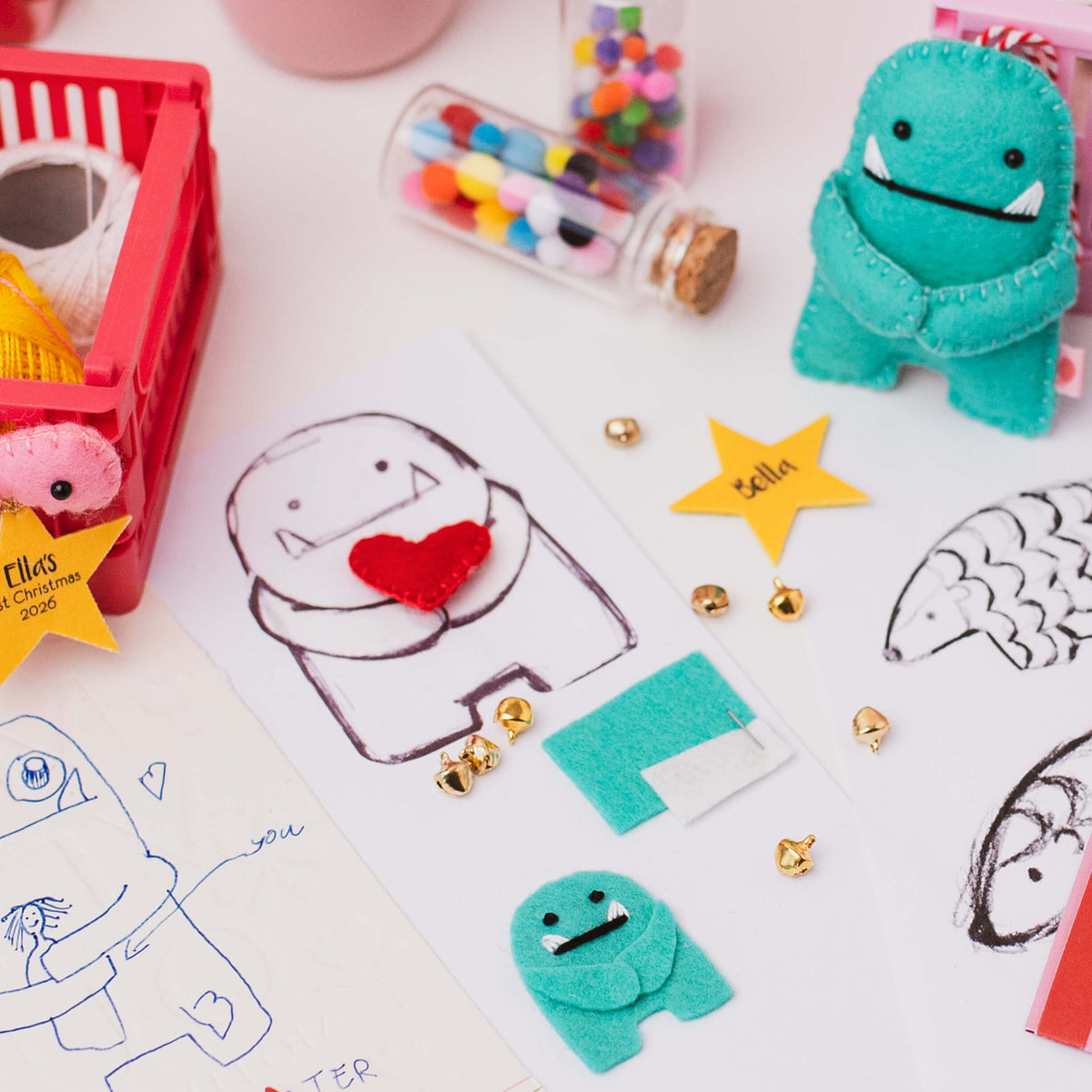 A styled flat-lay of handmade felt monster cornament on a white surface, including a teal felt monster, small gold jingle bells and drawing paper with c monster sketches.