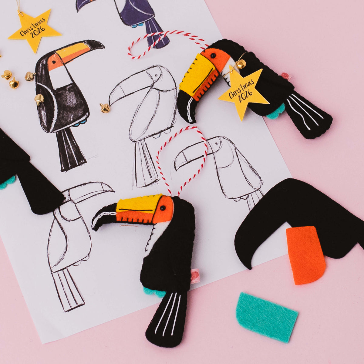 Felt Christmas toucan ornaments displayed on a pink background with a white sketch sheet of toucan sketches. The ornaments are made from black felt with bright orange, yellow, and teal accents, decorated with small gold bells and yellow star tags that read 'Christmas 2026'.