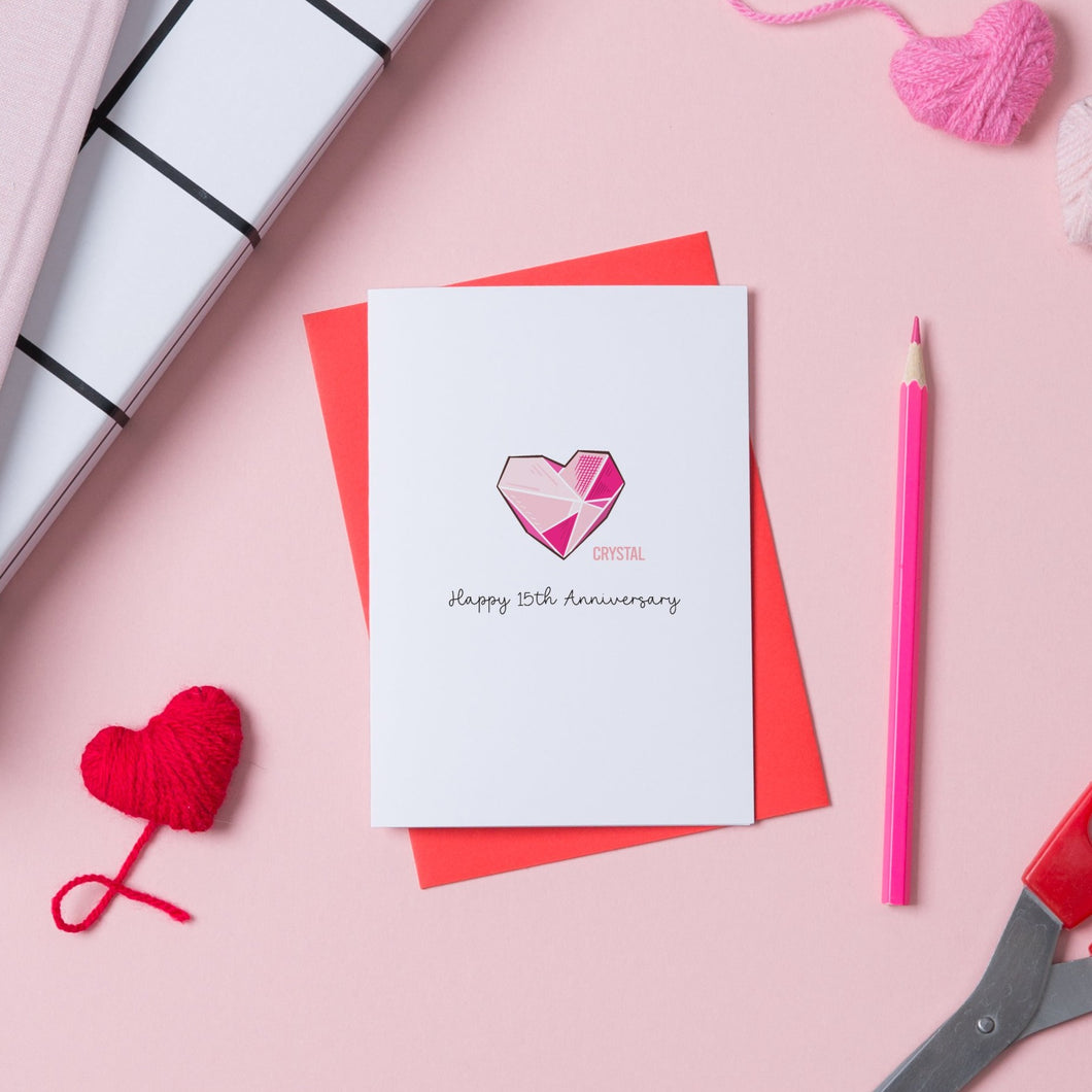 A 15th Wedding anniversary with a drawing of a crystal heart on the front and words saying Happy 15th Anniversary. The card sits on top of a red envelope.