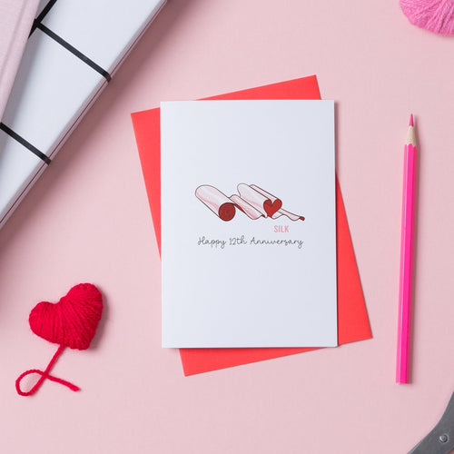 12th Wedding Anniversary card with an illustration of a roll of silk material draped into a heart shape. The card has Happy 12th Anniversary on the front and comes with a red envelope.