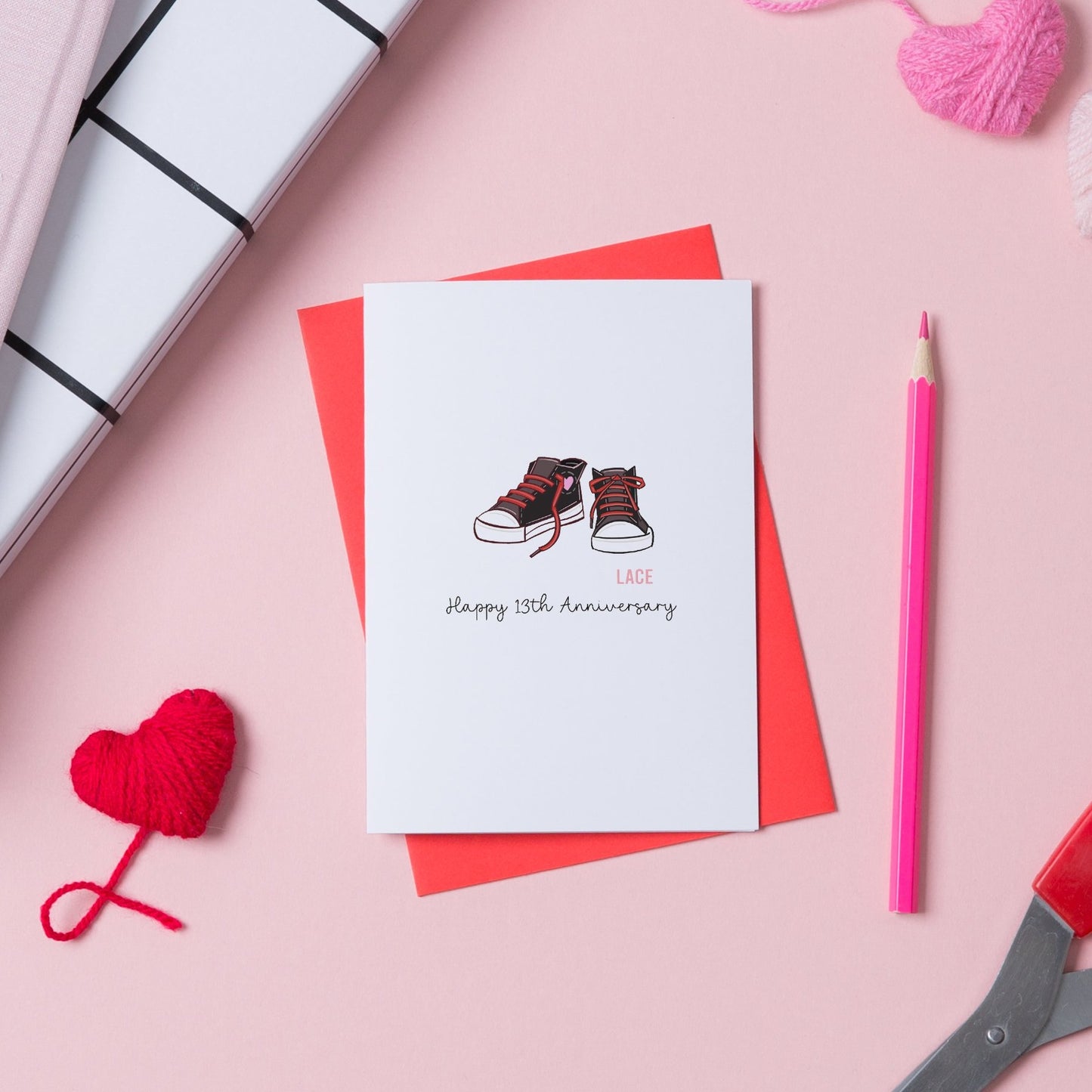 A picture of a card for a 13th anniversary. On the card is a pair of trainers with trailing laces, as the card is for a lace anniversary. There is a red envelope with the card.