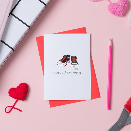A picture of a card for a 13th anniversary. On the card is a pair of trainers with trailing laces, as the card is for a lace anniversary. There is a red envelope with the card.