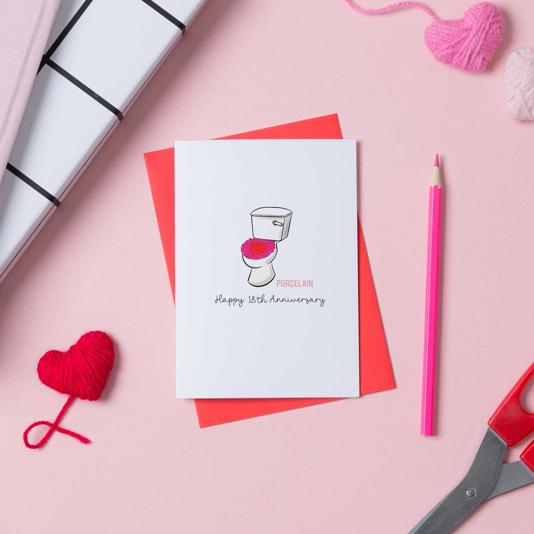 A white card with a picture of a porcelain toilet for an 18th anniversary card. the card sits on a red envelope on a pink background.