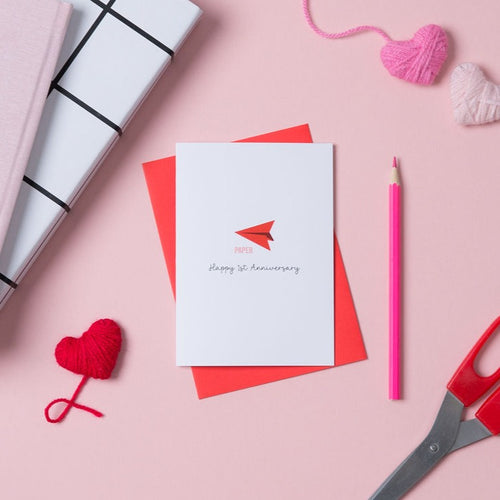 A white 1st anniversary card with a drawing of a red paper aeroplane on the front. The card is on top of a red envelope and next to it there is a pencil, hearts, books and a pair of scissors.